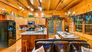 A cabin interior with wooden walls, a kitchen with granite countertops, a dining area with a table set for four, and a leather sofa with cushions. - 🏔🏔 180° Views 🏔🏔 ❸Decks ❸Masters ❷Game R♨Hot T (Sevierville)