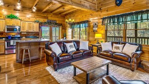 A cozy cabin interior with a wooden ceiling, a leather sofa, a wooden coffee table, and a kitchen area with a microwave and bar stools. - 🏔🏔 180° Views 🏔🏔 ❸Decks ❸Masters ❷Game R♨Hot T (Sevierville)