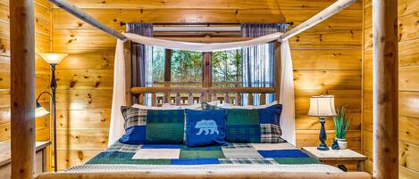 A wooden cabin bedroom with a four-poster bed, a window with curtains, and a nightstand with a lamp.