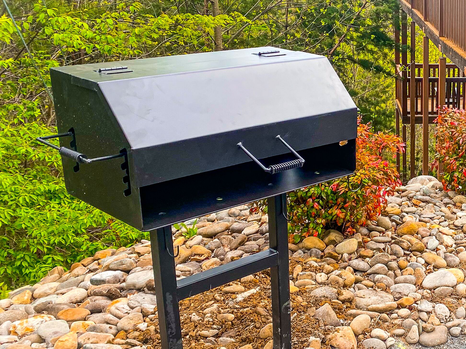 A black barbecue grill with a metal frame, set on a stone surface with greenery in the background.
