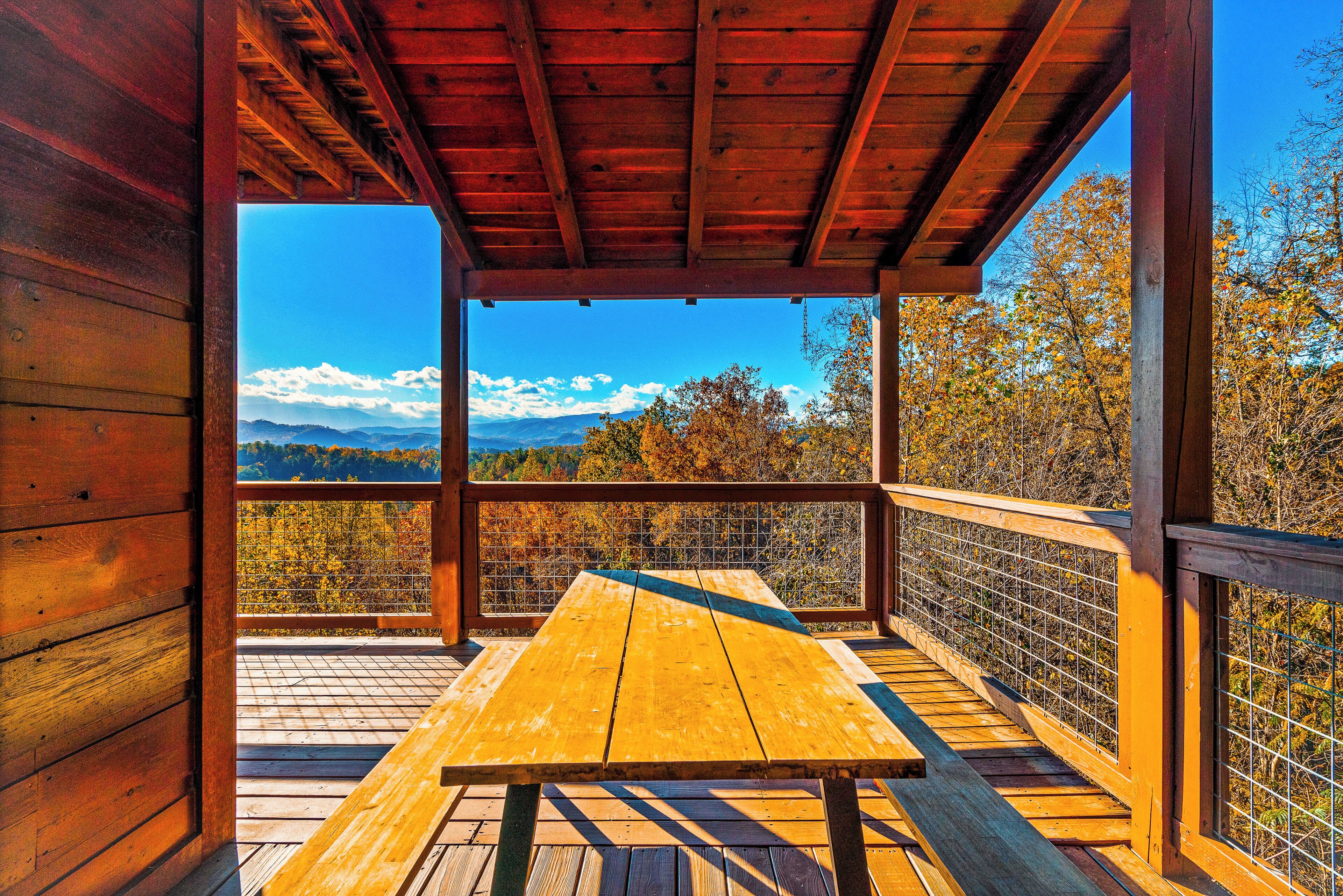A wooden deck with a picnic table, a wooden railing, and a view of mountains and autumn trees.