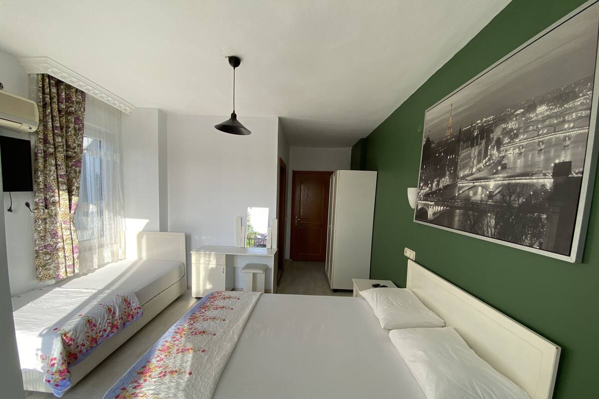 A hotel room with a large bed, a smaller bed, a desk, and a chair. There is a window with curtains, a wall-mounted air conditioner, and a picture of a cityscape.