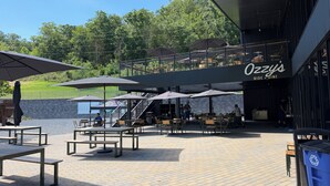 An outdoor seating area with tables and umbrellas, a covered stage, and a view of the surrounding greenery.