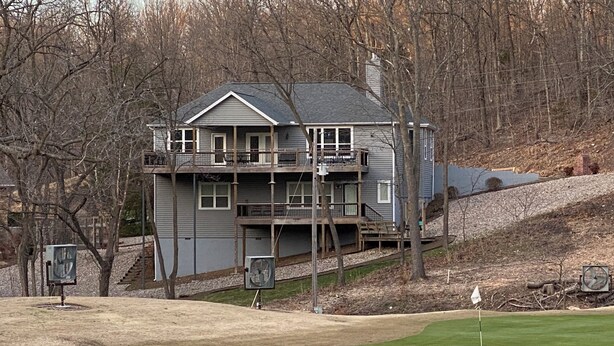 A two-story house with a balcony, a golf course in the foreground, and a wooded area in the background.