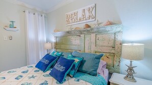 A bedroom with a bed featuring a sea-themed bedspread, a wooden headboard with a distressed finish, and a wall-mounted shelf with decorative items. - Captain's Cabana Gulf Front Condo (Galveston)