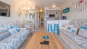 A cozy living room with two patterned sofas, a wooden coffee table, and a kitchen area with a white countertop and bar stools. - Captain's Cabana Gulf Front Condo (Galveston)