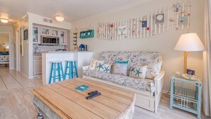 A cozy living room with a sofa, a coffee table, and a kitchen in the background. - Captain's Cabana Gulf Front Condo (Galveston)