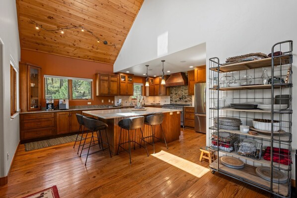 A modern kitchen with wooden cabinets, a large island with bar stools, and a metal shelving unit with dishes.