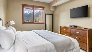 A bedroom with a bed, a dresser, a TV, and a window with a mountain view. - Alluring Mountain View Condo - The Heart Of Downtown! (Canmore)