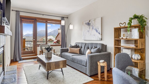 A modern living room with a grey sofa, a wooden coffee table, and a glass dining table. There is a wooden bookshelf with decorative items and a potted plant. A large window offers a view of mountains and a building outside. - Alluring Mountain View Condo - The Heart Of Downtown! (Canmore)