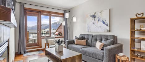 Una sala de estar moderna con un sofá gris, una mesa de centro de madera y vistas a montañas a través de una puerta corrediza de vidrio.