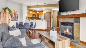 A modern living room with a fireplace, a flat-screen TV mounted above it, and a dining area with chairs and a table. - Alluring Mountain View Condo - The Heart Of Downtown! (Canmore)