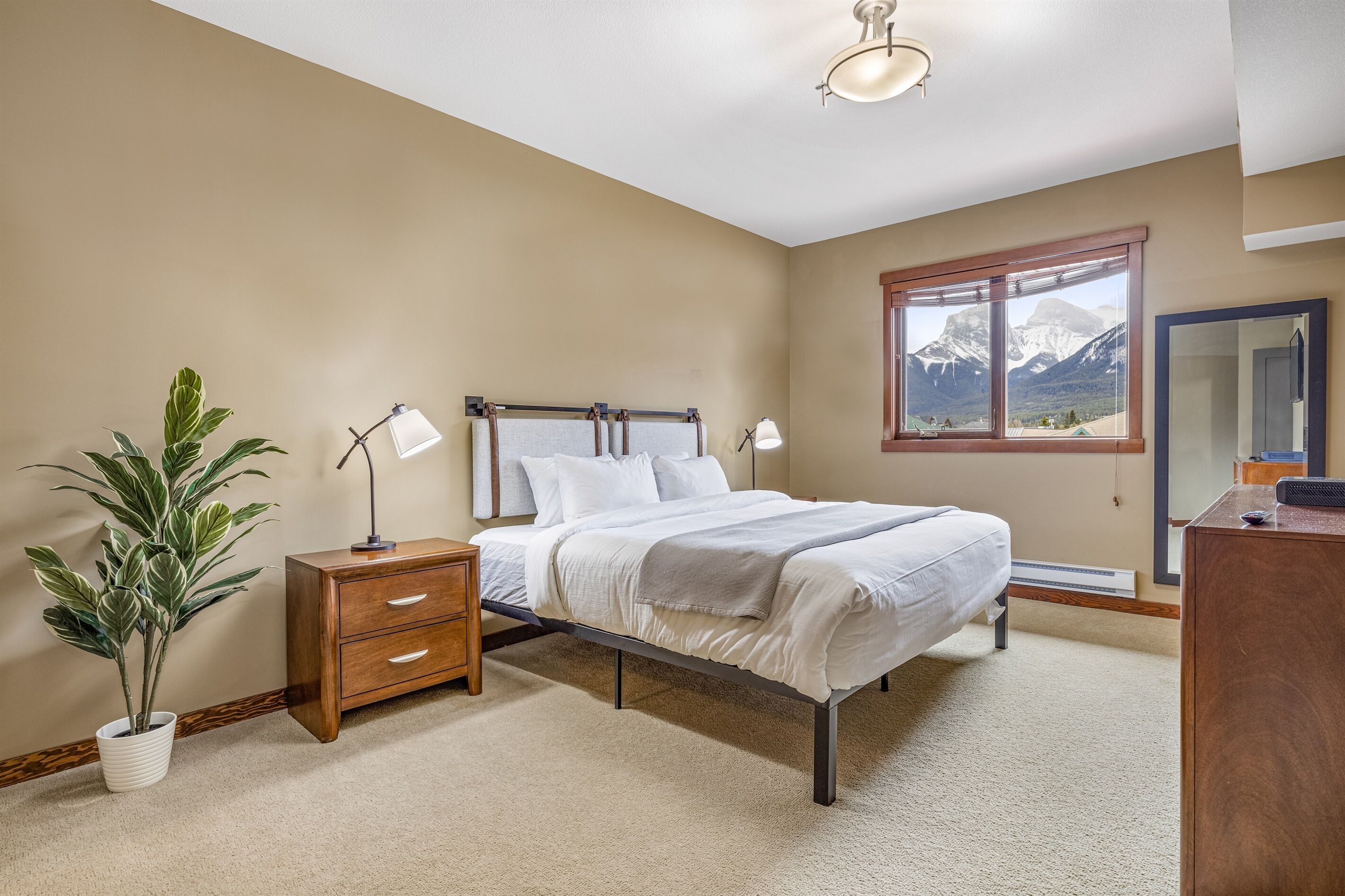 A bedroom with a bed, bedside lamps, a nightstand, a potted plant, and a window with a mountain view.