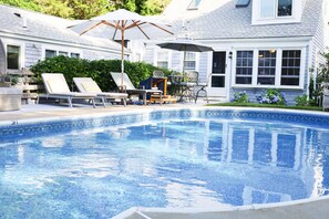 A swimming pool with lounge chairs and umbrellas, surrounded by a house with large windows and a garden. - Harbor Getaway Unit -1 (Harwich)