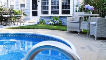 A residential backyard with a swimming pool, a patio with outdoor furniture, and a house with multiple windows and a covered porch.