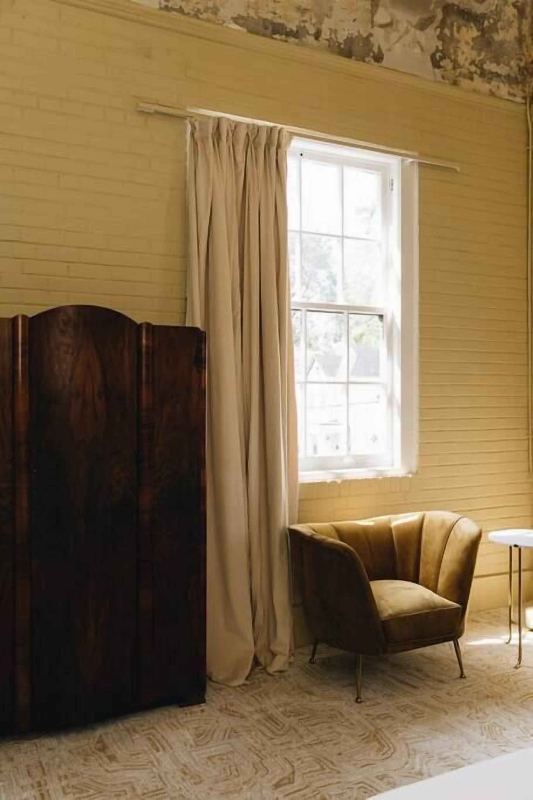 A room with a large wooden cabinet, a window with curtains, a brown armchair, and a small white table.