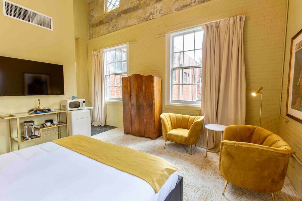 Une chambre d’hôtel comprenant un lit, une armoire en bois, deux fauteuils, une petite table, un téléviseur et une fenêtre avec des rideaux.