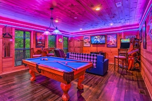 Une table de billard se trouve dans une pièce aux murs en bois et au plafond éclairé par des néons. Des affiches sont accrochées aux murs, il y a une télévision et une petite table avec des chaises.