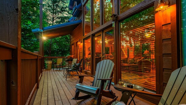Een houten terras met buitenmeubels, waaronder stoelen en tafels, en grote ramen met warme verlichting.