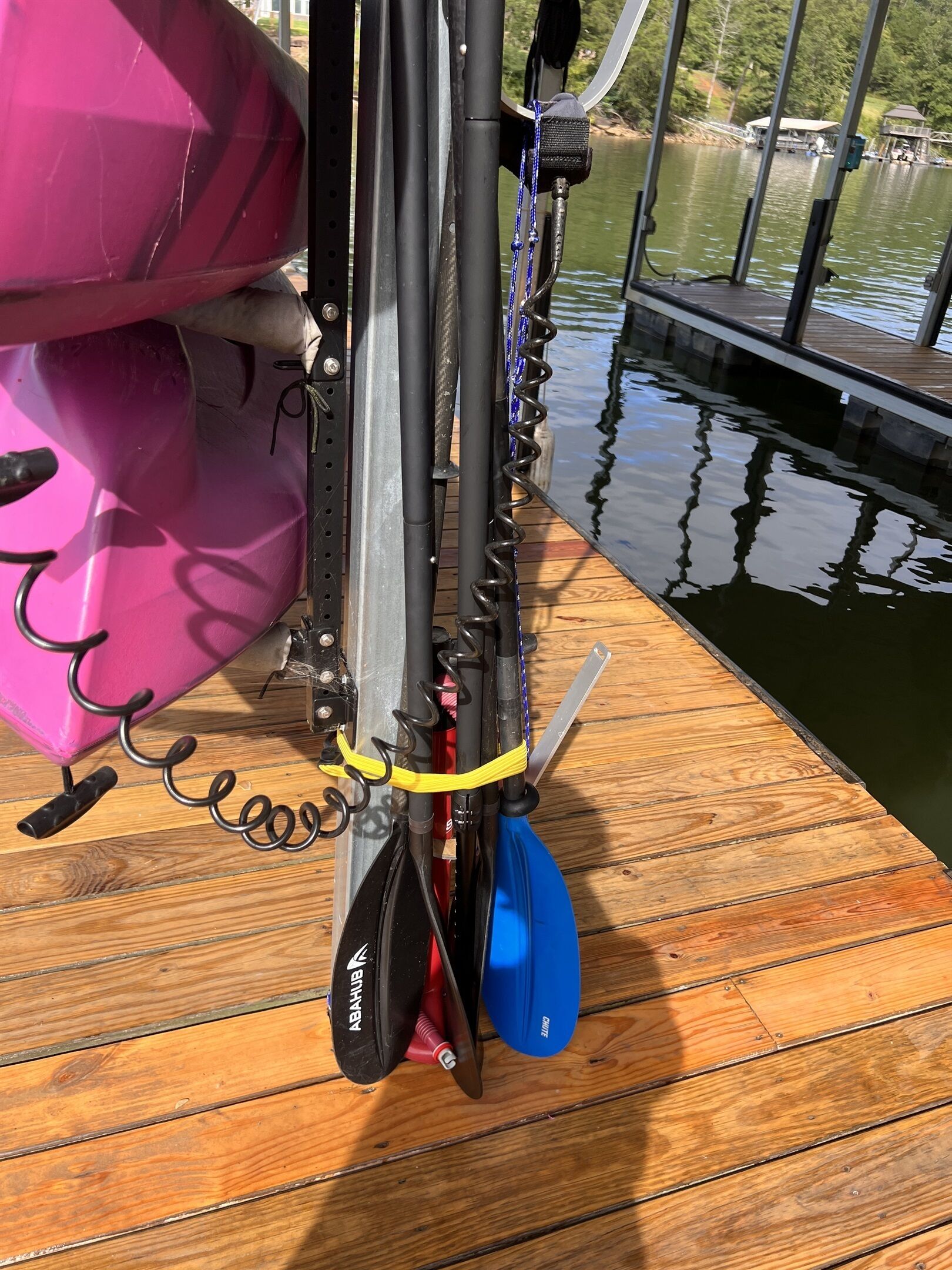 A kayak with a black and red paddle, a blue paddle, and a pink kayak bag on a wooden dock by a calm lake.