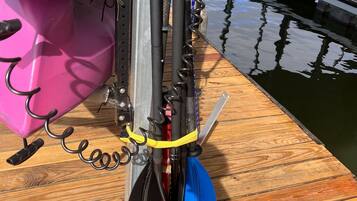 A kayak with a black and red paddle, a blue paddle, and a pink kayak bag on a wooden dock by a calm lake.