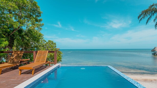 En klarblå pool med trädäckstolar, en träpergola och utsikt över havet.