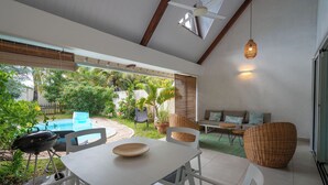 Un patio con mesa, sillas, una parrilla, piscina y un espacio de descanso con cojines.