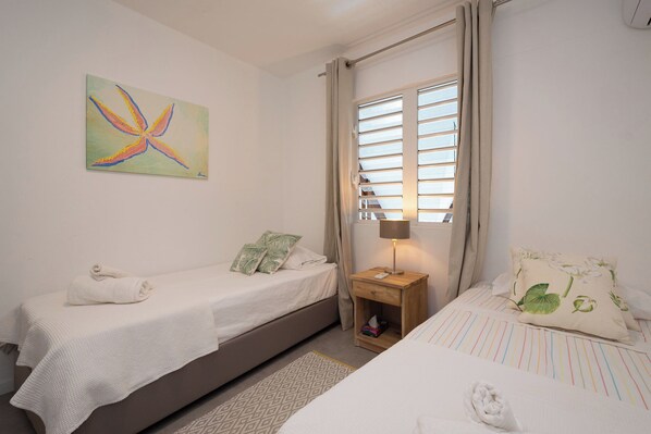 A bedroom with two single beds, a nightstand with a lamp, a window with curtains, and a colorful abstract painting on the wall.