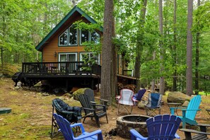 Une cabane dotée d’une terrasse, entourée d’arbres, et aménagée avec des chaises d’extérieur et un foyer.