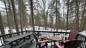 Une terrasse en bois avec des tables de pique-nique et des fauteuils Adirondack, entourée d’une forêt et d’un sol recouvert de neige.