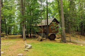 Une cabane entourée d’une forêt, avec un grand tonneau et des rochers au premier plan.