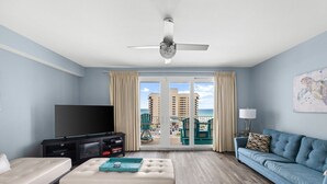 Una sala de estar con televisor, sofá, reposapiés y balcón con vista al océano y edificios.