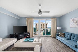 A living room with a TV, sofa, ottoman, and balcony with a view of the ocean and buildings.