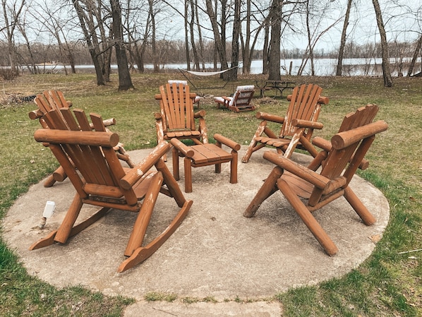 A circular arrangement of Adirondack chairs around a concrete table in a park setting with trees and a body of water in the background.