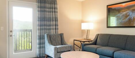 A hotel room with a sofa, armchair, small table, and a painting on the wall.