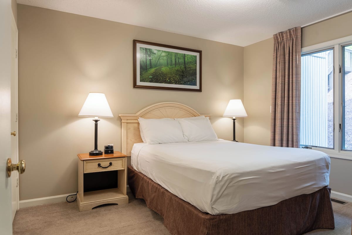 A bedroom with a bed, two bedside tables, a nightstand, a painting, and a window with curtains.