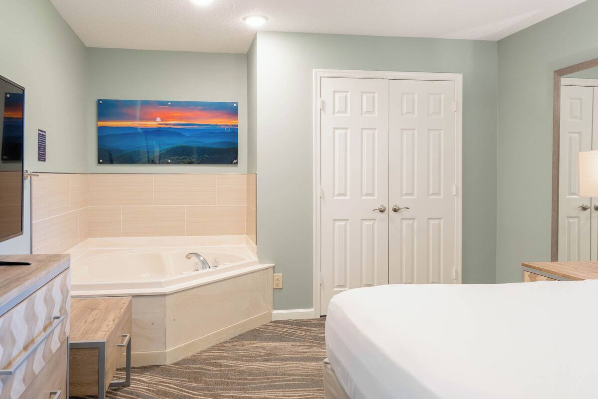 A hotel room with a bathtub, a bed, a door, and a painting on the wall.