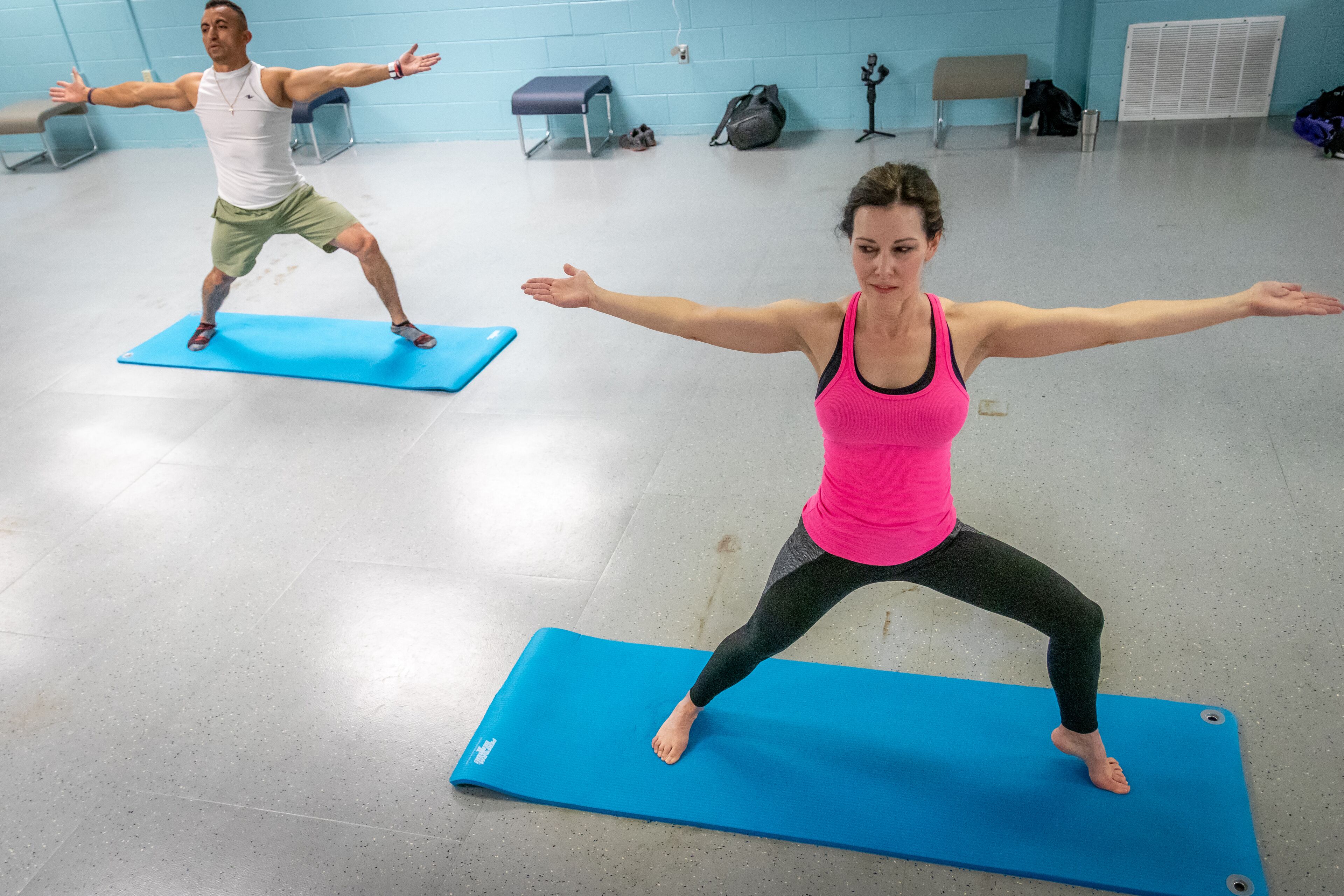 Two people practicing yoga on blue mats in a room with a tiled floor and a bench in the background.