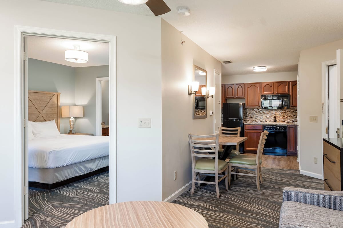 A hotel room with a bed, a dining table with chairs, a kitchen area with cabinets and appliances, and a hallway.