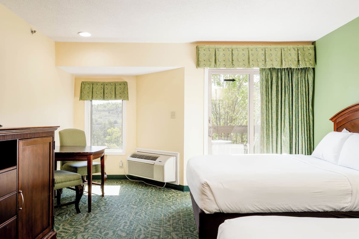 A hotel room with a bed, a desk, a chair, an air conditioning unit, and a window with curtains.