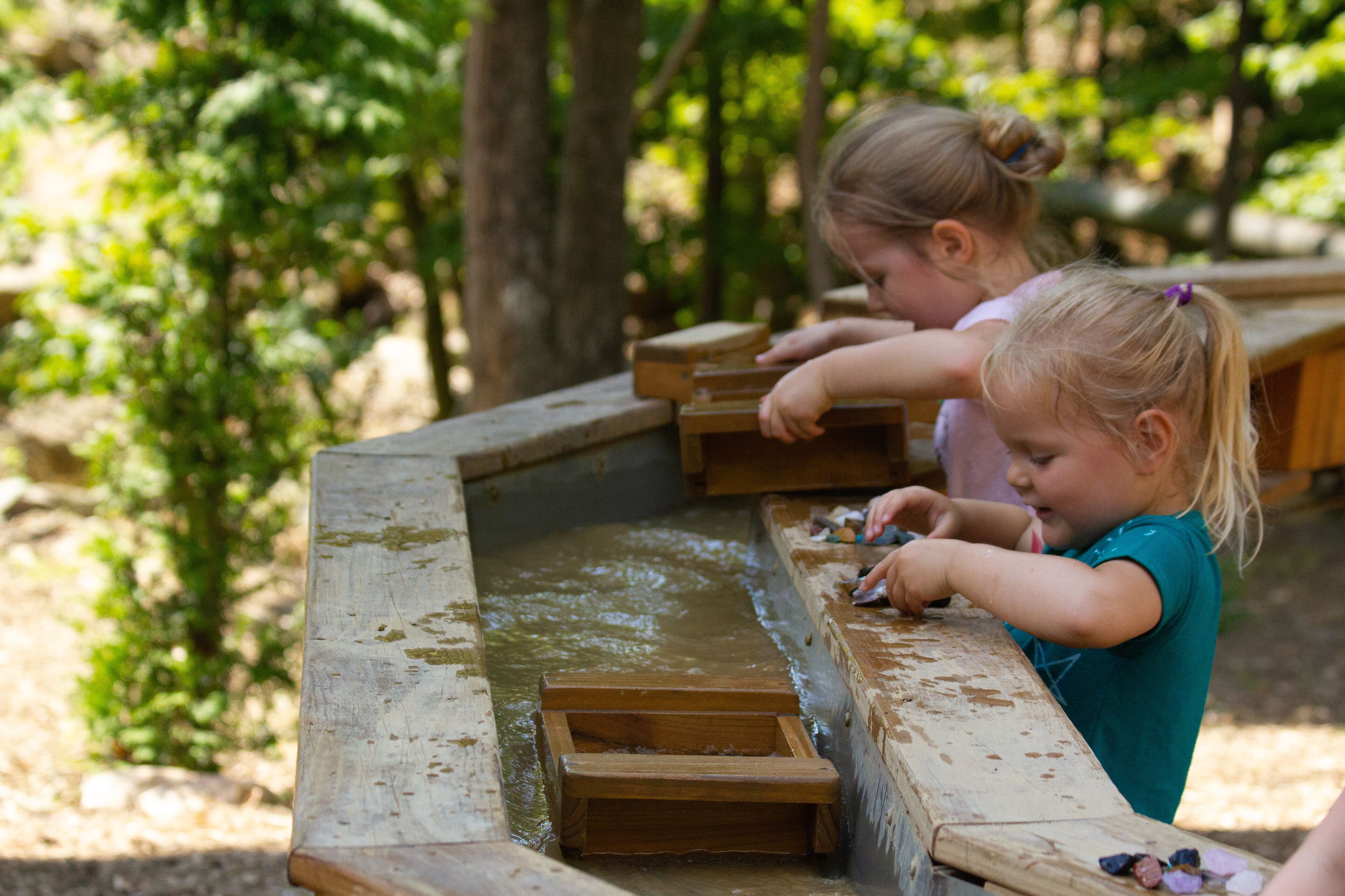 Two children playing in a wooden water feature surrounded by greenery.