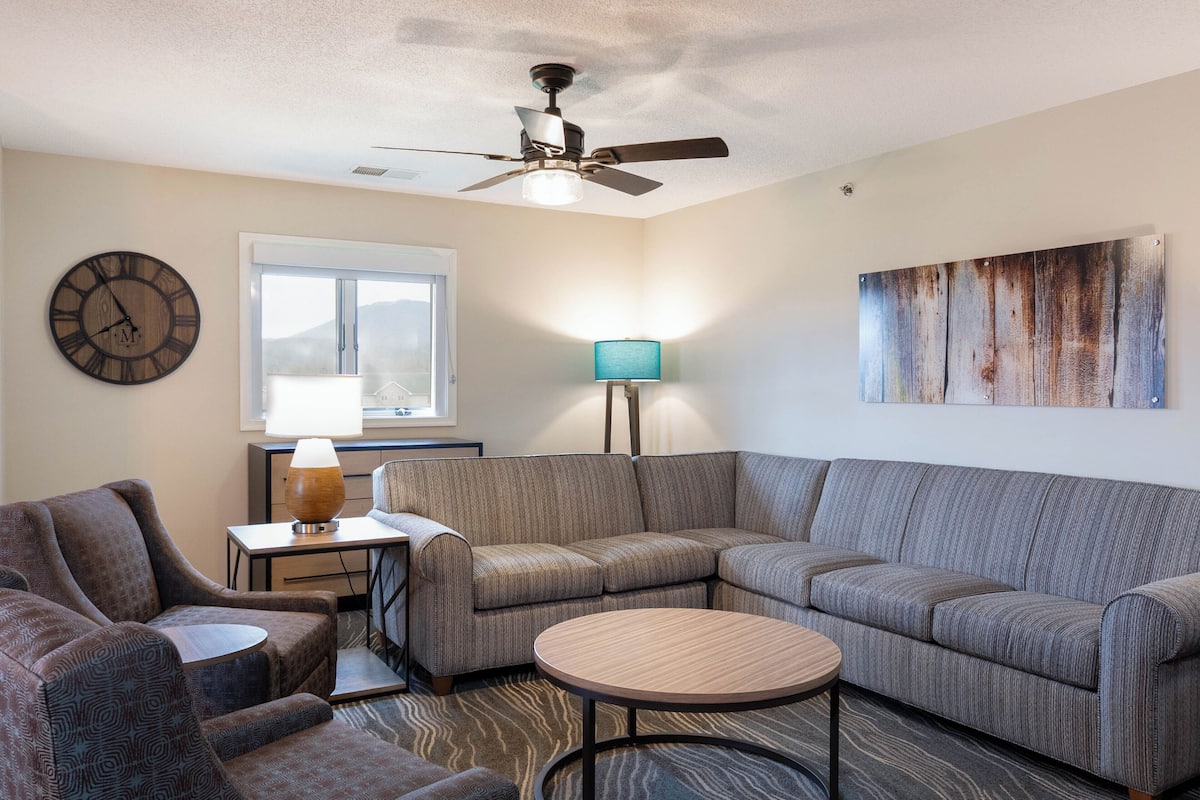 A living room with a sectional sofa, a round coffee table, a ceiling fan, a clock, a lamp, and a window with a view.