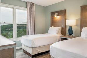 Habitación de hotel con dos camas, una mesita de noche con lámpara, un cabecero de madera y una ventana con cortinas.