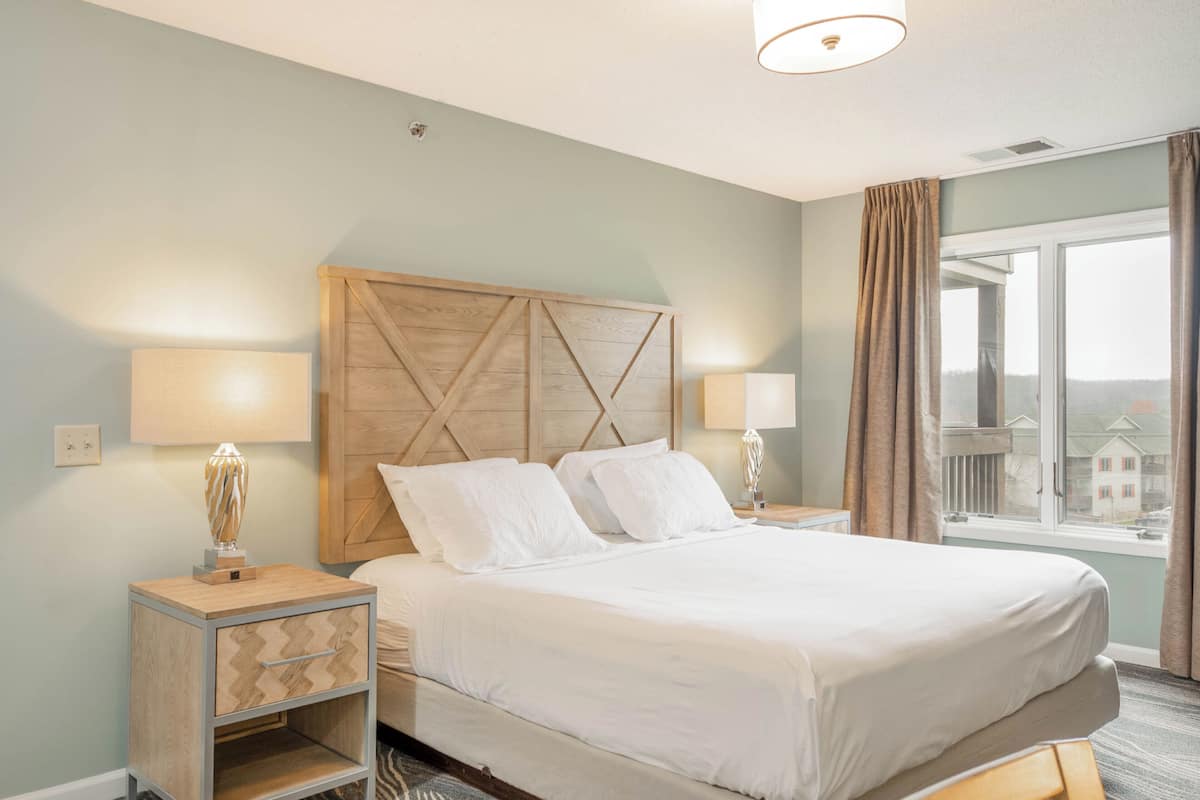 A hotel room with a bed, two bedside lamps, a nightstand with a lamp, and a window with curtains.