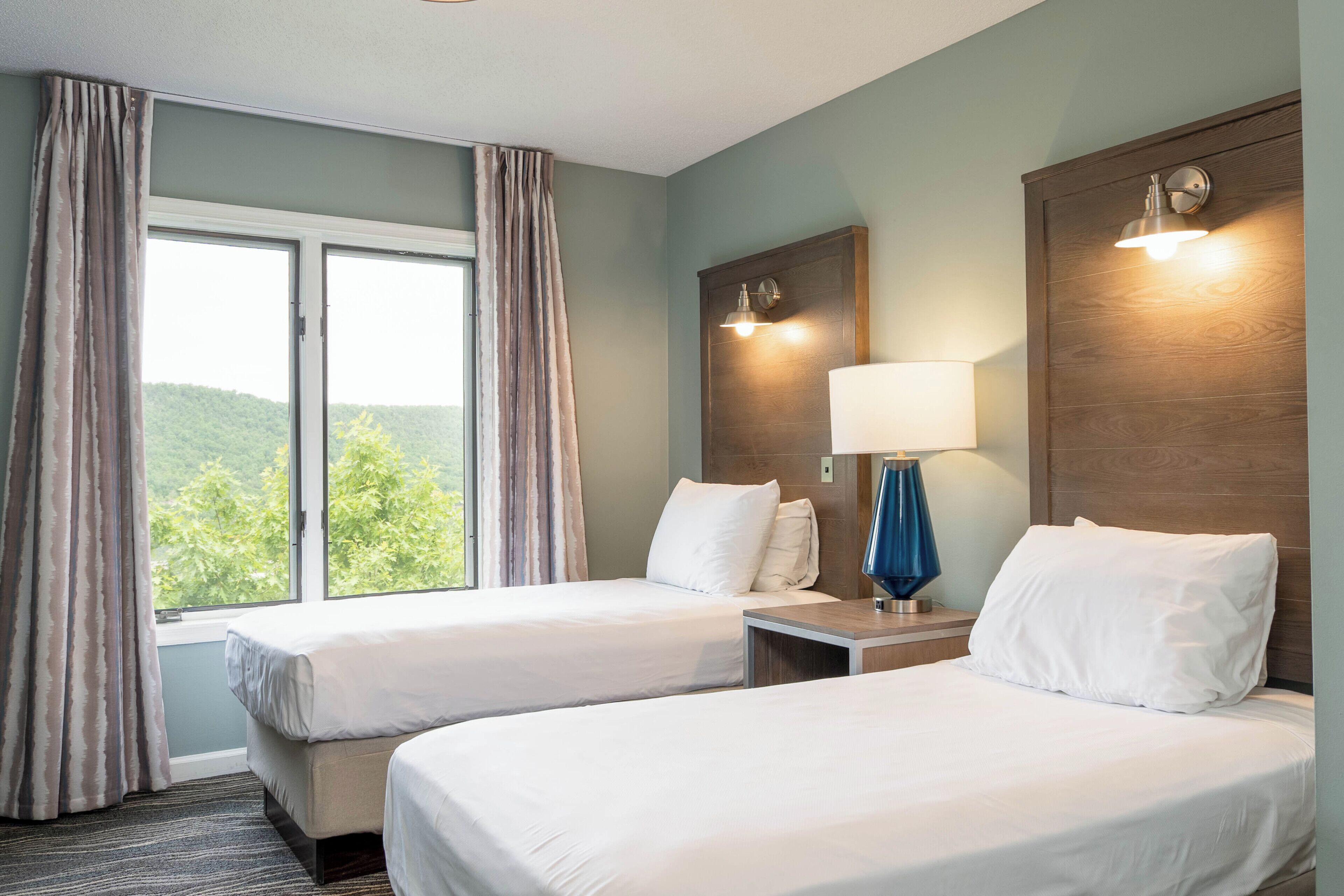 Een hotelkamer met twee bedden, een raam met gordijnen, een nachtkastje met een lamp en een wandlamp.