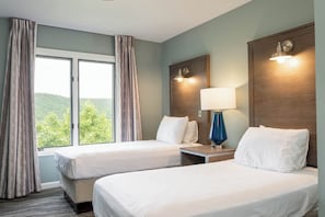 Habitación de hotel con dos camas, una ventana con cortinas, una mesita de noche con lámpara y un aplique de pared.