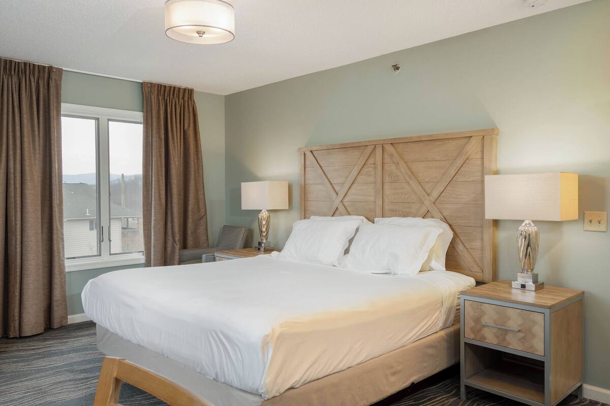 A hotel room with a large bed, two bedside tables, a wooden headboard, and a view through a window with curtains.