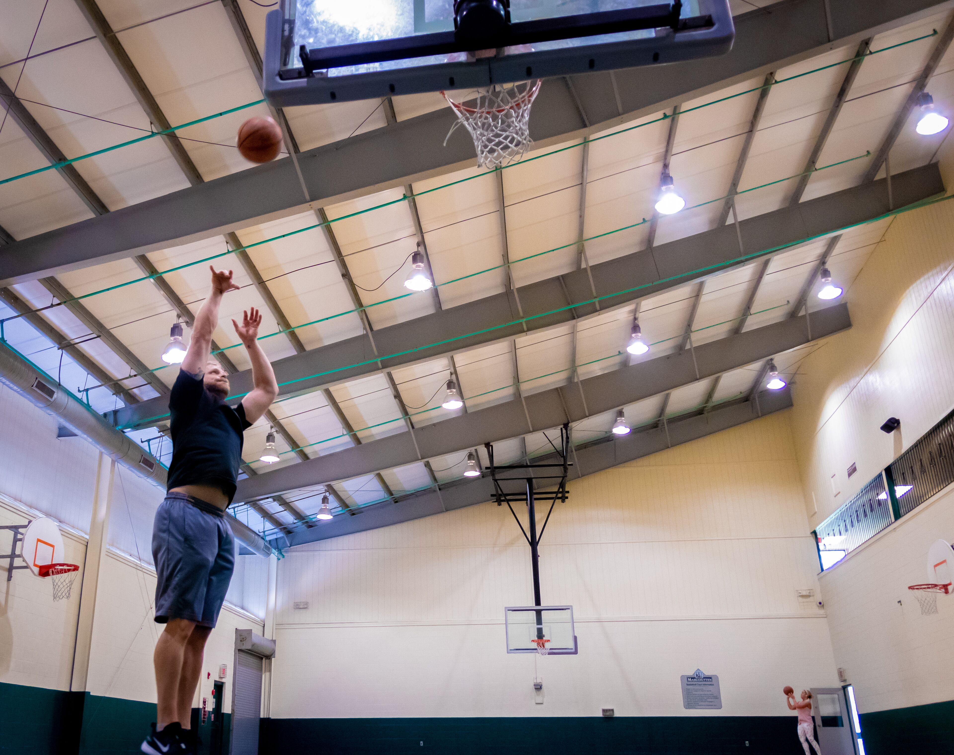 A basketball player in mid-air, aiming for a hoop in an indoor gymnasium with multiple basketball hoops and a high ceiling with visible lighting fixtures.