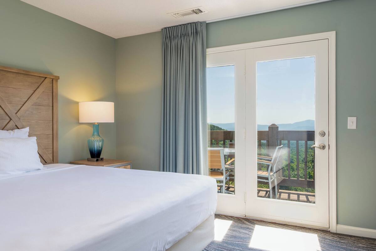 A hotel room with a bed, a nightstand, a lamp, a window with curtains, and a balcony with a view.