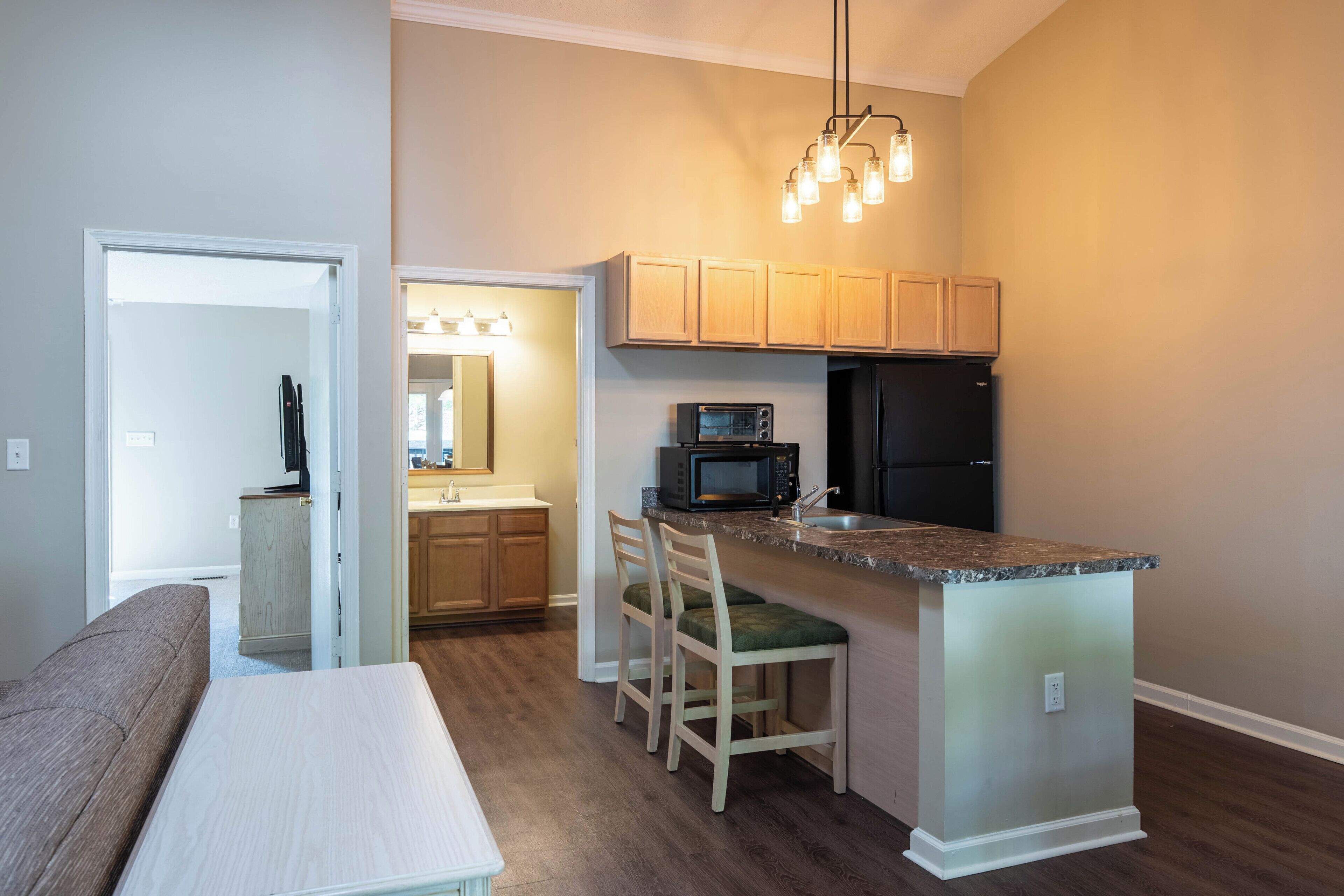A compact kitchen with wooden cabinets, a microwave, and a refrigerator. There is a bar area with stools and a bench.